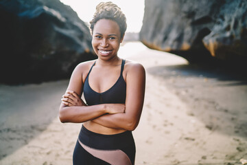 Portrait of cheerful African American young woman in tracksuit smiling at camera while crossed hands in front, joyful female jogger dressed in sportive top for workout posing at seashore in morning