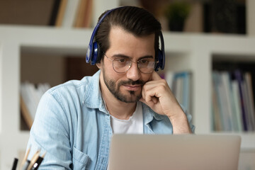 Wall Mural - Close up focused man in glasses and headphones using laptop, looking at computer screen, watching webinar or training, studying online, student listening to lecture, involved in internet lesson