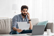 © Prostock-studio - Arab freelancer man working remotely with smartphone, laptop and documents, sitting on sofa at home