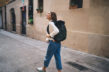 Wall Mural - Caucasian female tourist with travel backpack walking around historic quarter enjoying solo vacations, casual dressed woman 30 years old exploring city streets during international excursion in town