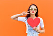 Beautiful smiling woman holding a red heart over orange background. Fashion portrait stylish pretty woman in sunglasses outdoor.
