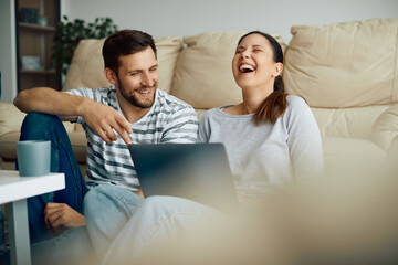 Wall Mural - Cheerful couple laughing while using laptop and reading something funny on the Internet at home.