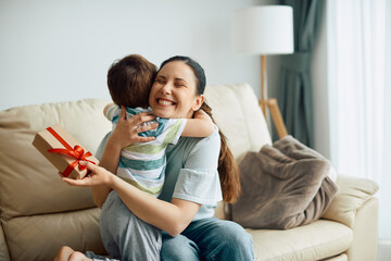 Wall Mural - Happy mother embracing her son while receiving a gift box from him at home.