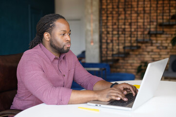 Smart and confident African-American freelancer is using laptop for work on the distance, black guy sitting at the desk and typing, develods software, sending emails to business partners
