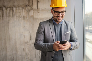 Smiling businessman with helmet on head standing next to a window in building in construction process and using phone.