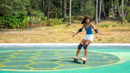 Attractive Asian woman with safety skateboarding knee pad skating at skateboard park by the beach. Happy female enjoy summer outdoor active lifestyle play extreme sport surf skate at public park.