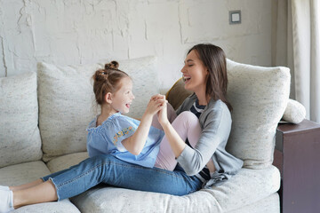 Cute little kid daughter laughing playing with mother on sofa, happy mother relaxing having fun with funny small child girl bonding enjoying leisure together in living room.