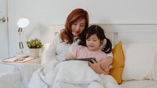Asian Grandmother Using Tablet Read Fairy Tales To Granddaughter At Home. Senior Chinese, Grandma Happy Relax With Young Girl Before Bedtime Lying On Bed In Bedroom At Home At Night Concept.