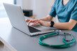© dikushin - Close-up hands of unrecognizable female physician in medical uniform working typing on laptop keyboard sitting at desk on background of window in dark office room.