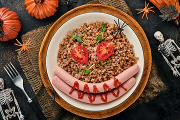 Halloween food art idea healthy lunch for kids. Boiled buckwheat porridge, meat sausages, tomato and microgreens of peas in a white plate for kids breakfast or lunch on stone background. Top view.