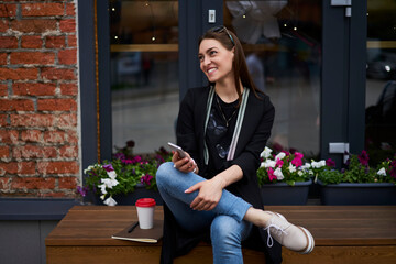 Wall Mural - Cheerful hipster girl in casual clothes laughing while creating publication text sitting outdoors in city, happy Caucasian student with modern mobile phone looking away and smiling during daytime