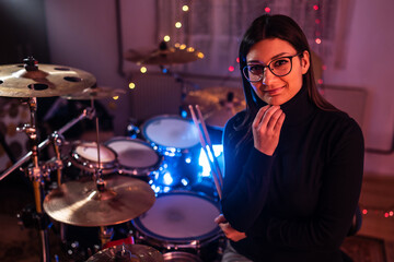 Front view portrait of adult caucasian woman posing in front of drums set in dark room or studio - Drummer woman standing looking to the side at night - copy space real people