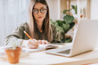 © Anna - Thinking Freelance woman in eye glasses with pencil write in note book, typing at laptop and working from home office. Happy girl on workplace at the desk. Distance learning online education.