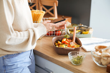 Wall Mural - Young woman taking photo of baking dish with vegetables in kitchen