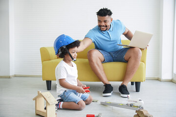 Little cute African engineer boy with helmet playing toys like plane, wooden car, hand tools on the floor while father sitting on sofa working with laptop look and pat head his son at home