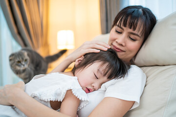 Asian mother hugging daughter sleeping on her shoulder in living room.