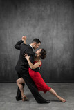 Couple of professional dancers in elegant suit and red dress in a tango dancing movement on dark background. Handsome man and woman dance looking  eye to eye.