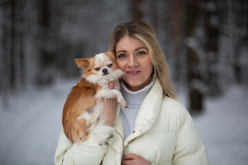 Blonde young female holding ginger and white chihuahua in her hands. Snow and trees on the background