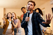 © Kostiantyn - Love singing. Enthusiastic handsome young guy singing with microphone while playing karaoke with friends at home, standing in the modern apartment