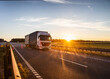 A white truck with a semitrailer carries cargo along the highway against the backdrop of a sunny sunset in the evening. Business and freight concept, copy space for text