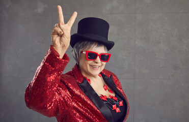 Smiling active grandma in the mood to party. Happy positive funny retired senior woman in black top hat, cool glasses, jewelry and bling sequin concert outfit doing V sign on gray studio background