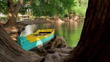 Bright Boat Tied To Tree Roots Floating On Rippling Water Of Calm Camecuaro Lake On Sunny Day In Michoacan, Mexico