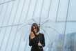 © Mikel Taboada/Westend61 - Smiling woman using mobile phone while holding umbrella against glass building