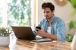© steve brookland/Westend61 - Serious male freelance worker holding mobile phone while using laptop at table