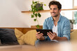 © steve brookland/Westend61 - Smiling handsome man holding digital tablet while sitting on couch at home