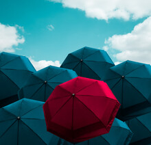 Prominent Vivid Red Umbrella Among Group Of Blue Parasol On Cloudy Blue Sky, Protection Item For Sunny And Rainy Weather. Protective Idea With Repetition Pattern. Differentiate, Outstanding Concept.