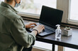 © Burak Kavakci - young woman sitting in a mask at coffee shop working on laptop, using disinfectant