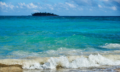 Wall Mural - Marine wave breaking on the shore of a sandy beach with a small island with a palm tree in the background on the island of San Andres in the Colombian Caribbean.