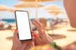 © Celt Studio - mockup image. woman use cell phone on beach, summer holiday. woman holding mobile phone with blank desktop screen on the beach. Mobile phone in female hand on the background of the resort beach