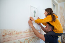 Happy Daughter And Father Hanging Picture On Wall. Medium Shot Of Caucasian Girl Sitting On Parents Neck And Holding Paint Frame During Apartment Decoration. Spending Time Together, Family Concept