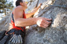 Climber In Red T-shirt Climbs A Gray Rock. A Strong Hand Grabbed The Lead, Selective Focus. Strength And Endurance, Climbing Equipment: Rope, Harness, Chalk, Chalk Bag, Carabiners, Braces, Quickdraws