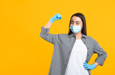 Woman with protective mask and gloves showing muscles on yellow background, space for text. Strong immunity concept