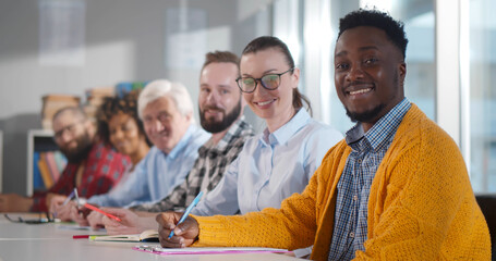 Multiethnic adult people sitting in row at desk and smiling at camera in classroom