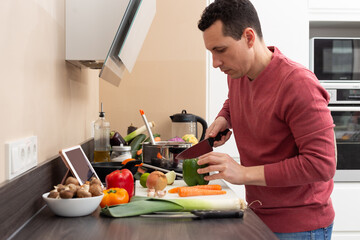 Man wearing an apron cooking at home in a modern kitchen vegetarian and vegan food looking at a tablet. Healthy recipe.