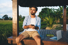 Positive young guy with laptop using smartphone in picturesque field