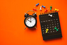 Black Analog Alarm Clock, Opened Calendar And Multi Color Thumbtacks On Grunge Orange Paper Background