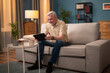 © ABCreative - A contented senior talks to his children, grandchildren over the internet. Senior relaxes on the sofa playing games on the tablet. Elegant elderly man is relaxing on the sofa surfing the internet