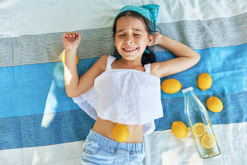 View from above of a little girl smiling with closed eyes and lying on the blanket with lemons in the park. Pretty kid smiling and having summer picnic with family on a sunny day outdoors.