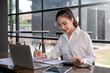 © amnaj - Asian businesswoman calculating accounting numbers with a laptop and taking notes at the office.
