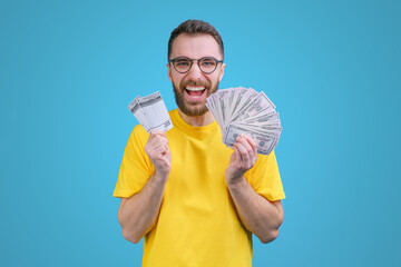 Wall Mural - Portrait of happy euphoric bearded man in yellow t-shirt posing with lottery ticket and ward of dollar bancknotes in hands as betting, gambling, money win concept