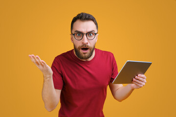 Young bearded man in trendy hipster eyeglasses holding tablet computer and looking directly at the camera with confused face expression