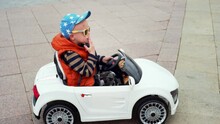 4 Years Old Caucasian Boy In Sunglasses Is Driving A White Toy Battery Car At The Street. Fancy Kid In Electric Car