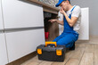 © yaroslav1986 - Handyman in overalls looks at the pipes under the sink in the kitchen and shows that there is an unpleasant smell, there is a box of tools next to it