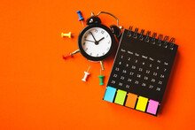 Black Analog Alarm Clock , Opened Calendar With Paper Tag And Multi Color Thumbtacks On Grunge  Orange Paper Background, Top View