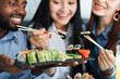 © sofiko14 - Lunch at a Asian restaurant. Close up shot of happy grinning multiethnic friends, black man and two pretty girls, eating sushi rolls with chinese sticks. Focus on plate with rolls and sticks