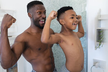 African American Father And Son In Bathroom, Looking In Mirror Showing Muscles